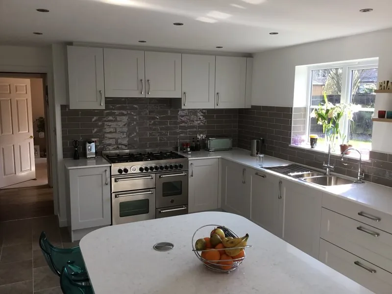 A fitted kitchen with pale shaker cabinets, a range cooker, grey tiled splashback and a white worktop island